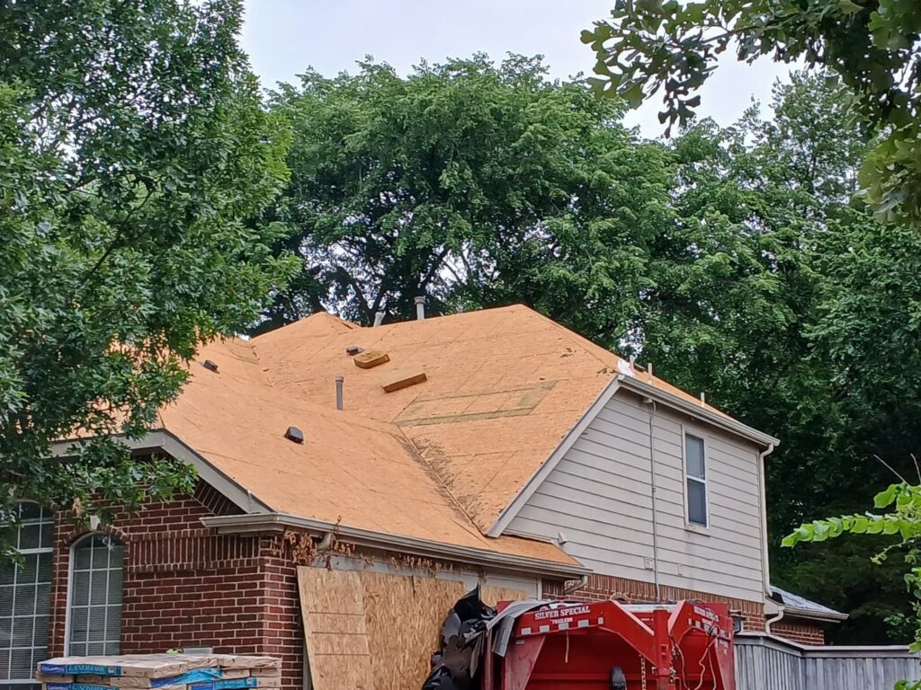 The image shows a residential house with its roof stripped down to the decking and underlayment, indicating a re-roofing project in progress. A red dumpster and bundles of new asphalt shingles are visible on the ground.