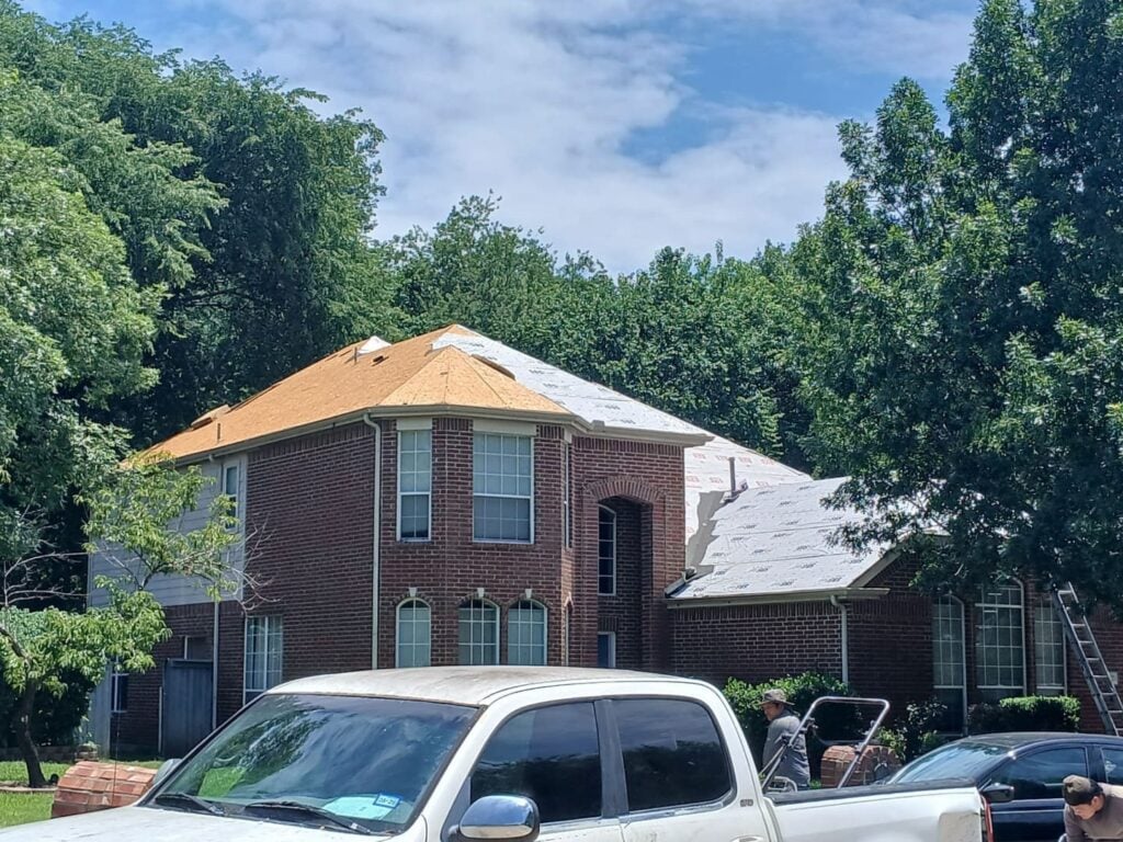 A brick house with a roof partially stripped to the decking and partially covered with synthetic underlayment, indicating active roofing work. Two workers are visible near a white pickup truck in the foreground.