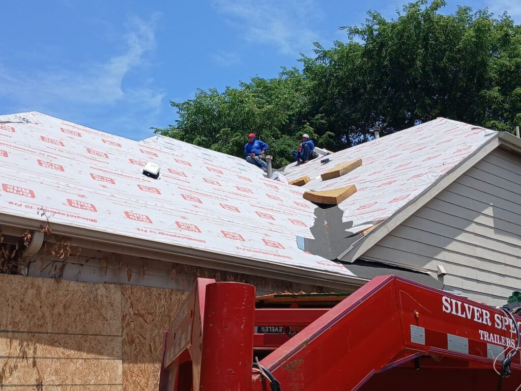 Two roofers are actively working on a residential roof, which is currently covered with ShingleLayment-15 Pro underlayment. A red 'Silver Spec Trailers' trailer is visible in the foreground.