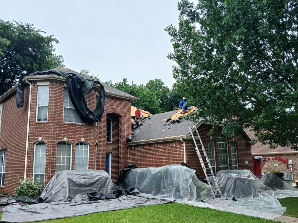 An exterior shot of a brick house where roofers are actively stripping and replacing the roof. Protective tarps cover the landscaping and ground around the house, and a debris chute is visible on the right.
