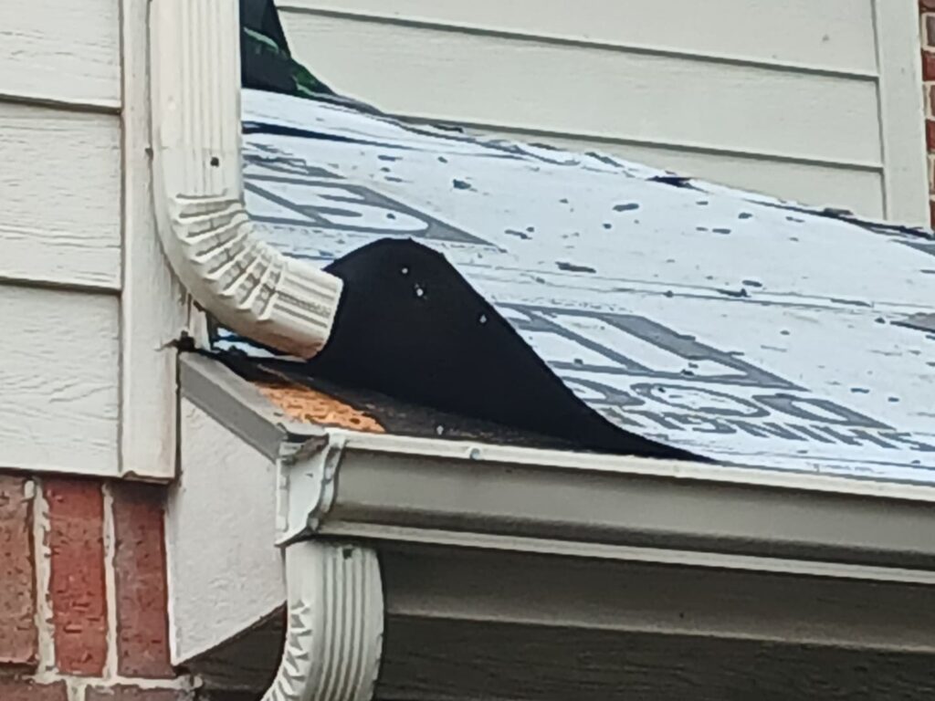 A close-up view of a residential roof showing exposed underlayment, a white gutter, and a section of the house wall with siding and brick.