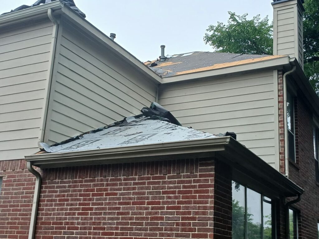 An image of a residential house with a partially removed asphalt shingle roof, exposing the underlayment and some bare decking. Gutters and siding are also visible.