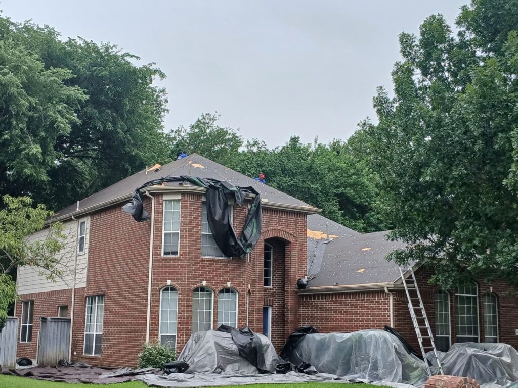 A large brick house with its roof partially removed, showing underlayment and exposed decking. Tarps cover landscaping and debris on the ground, and two workers are visible on the roof.