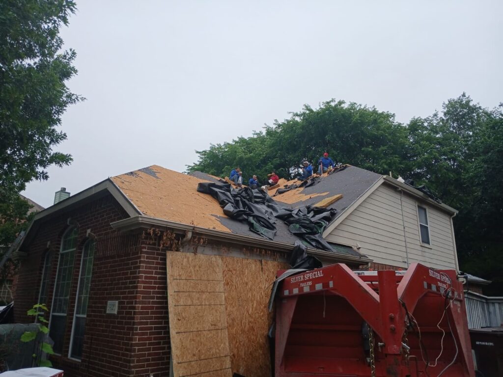 A crew of roofers is actively working on a residential roof, which is partially stripped of old material, revealing the decking and some underlayment. A large red dump trailer is parked in front of the house.