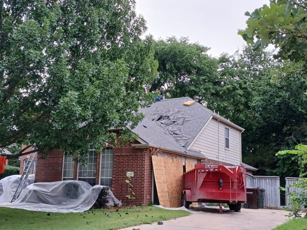 A residential house with a red dump trailer in the driveway, showing a roof replacement in progress with old asphalt shingles being removed. A worker is visible on the roof, and the front yard is covered with tarps.