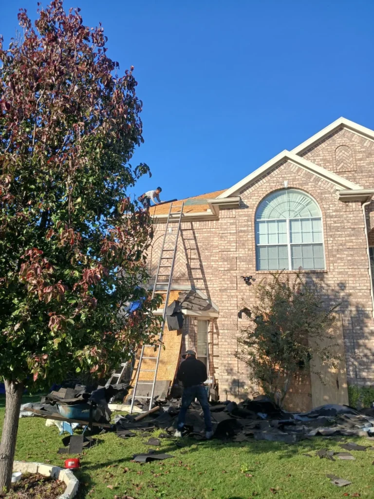 This image captures a residential roof replacement in progress, with two crew members on the roof and ladder, and another on the ground amidst removed asphalt shingles. The house is a two-story brick structure with a prominent arched window.