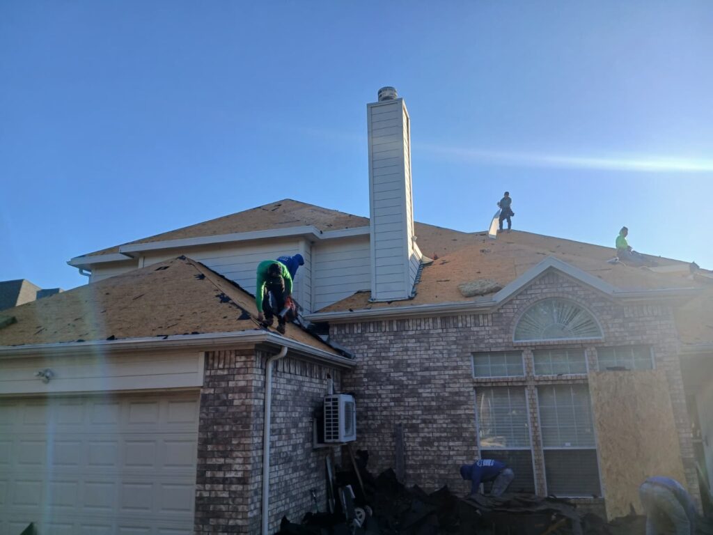 Several roofers are actively working on a residential house, tearing off old asphalt shingles from the roof deck under a clear blue sky. A pile of removed roofing debris is visible on the ground next to the house.