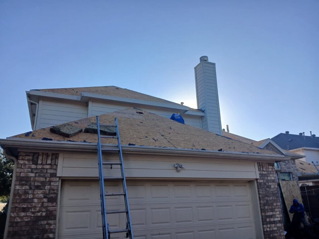 A residential roof is shown partially stripped, exposing the plywood decking and black underlayment. A ladder leans against the house, and the sun is low in the sky behind the structure, creating a backlit effect.