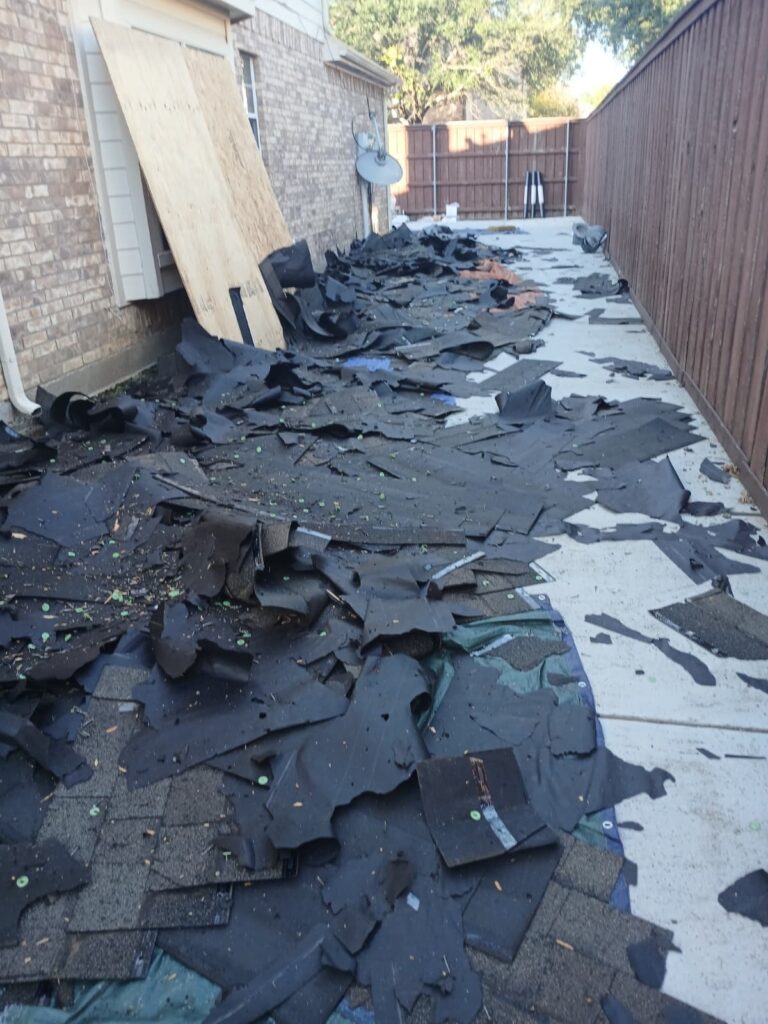 A narrow side yard is completely covered with discarded asphalt shingles and underlayment, indicating a roof tear-off in progress. A large sheet of plywood leans against the brick wall of the house.