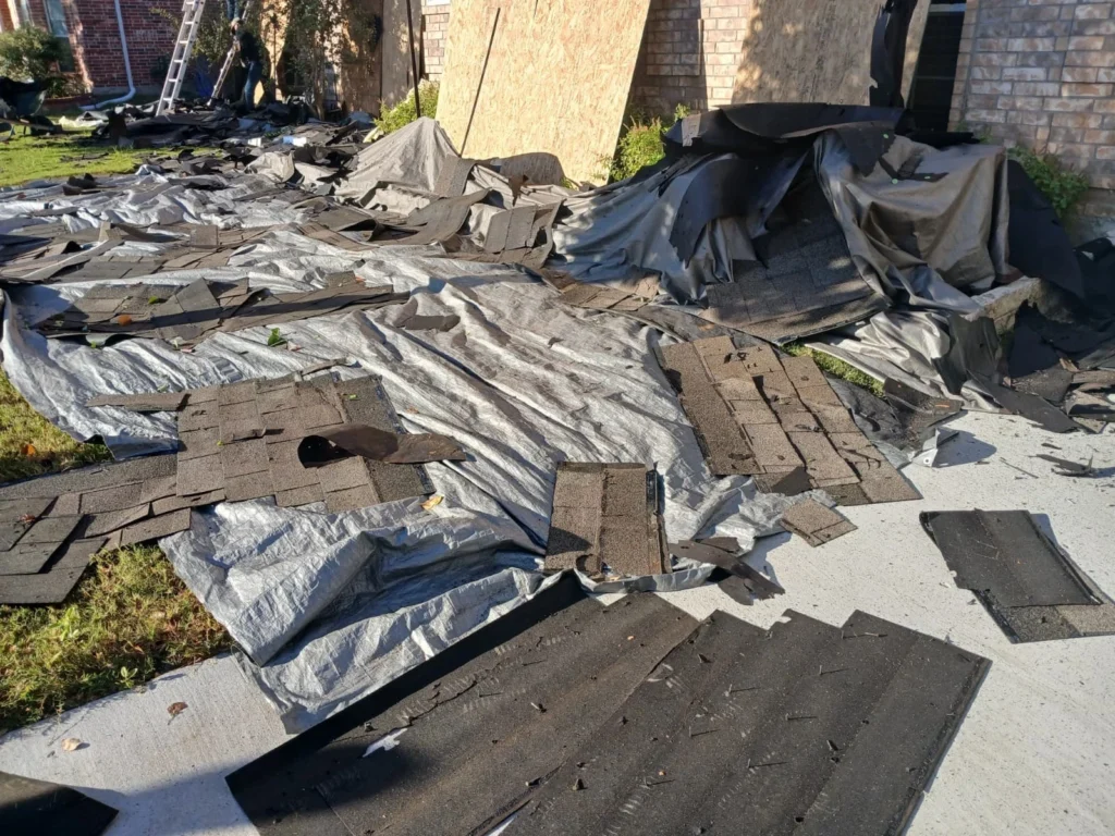 A residential yard is covered with tarps and piles of torn-off asphalt shingles, indicating a roof replacement project in progress. A ladder and a worker are visible in the background near the house.