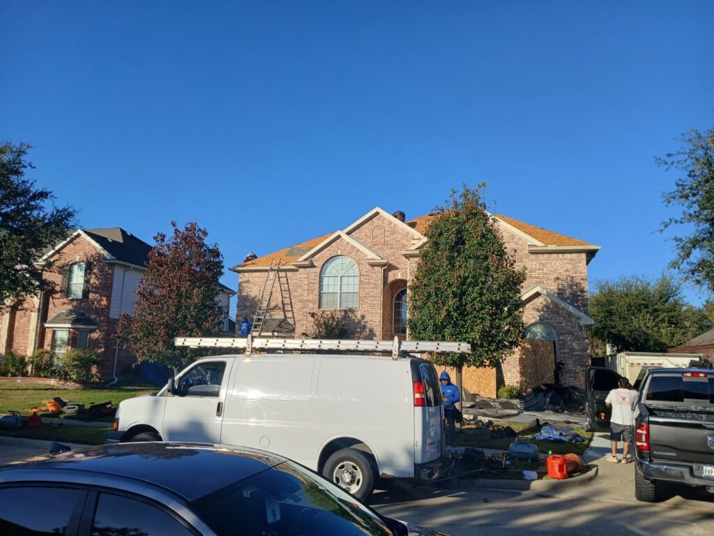 A residential house is undergoing roof replacement, with workers on the roof and ground, and a work van and pickup truck parked in front. Debris from the old roof is scattered on the lawn and driveway.