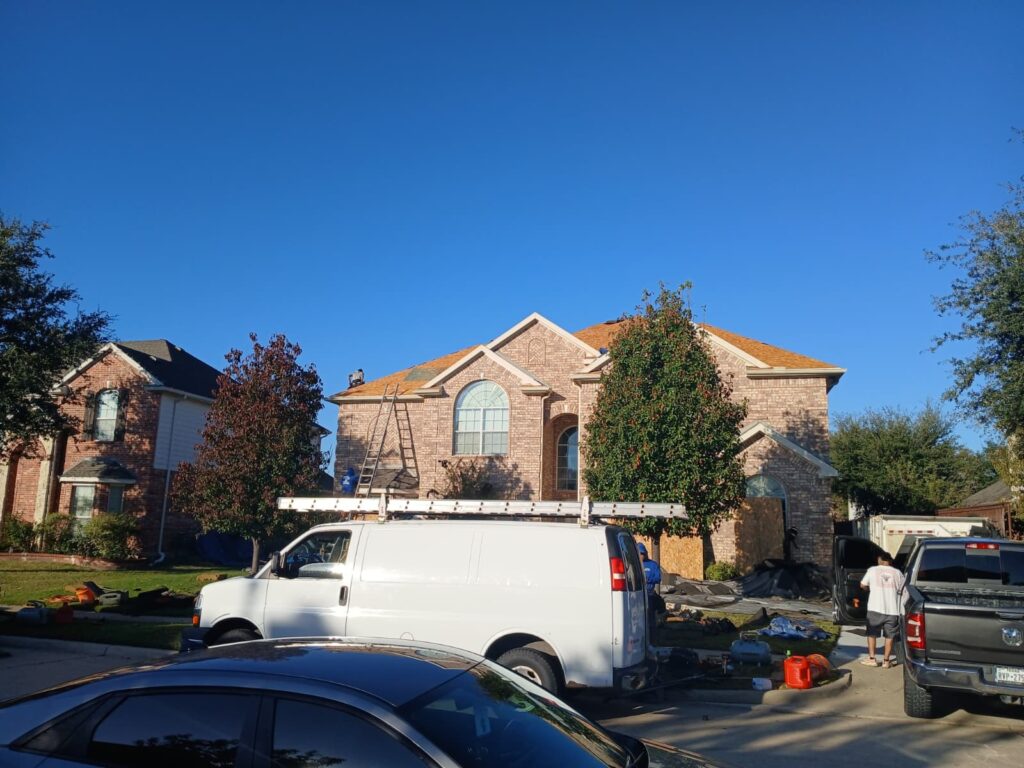 A residential house with a new roof being installed, showing workers on the roof and ground, a white work van, and a dark pickup truck. Roofing debris and materials are spread on the lawn.