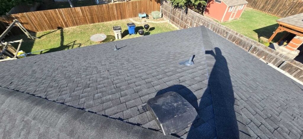 An aerial view of a newly installed dark gray asphalt shingle roof, showing the ridge line, a pipe boot, and a vent. The backyard with a wooden fence, shed, and various outdoor items is visible below.