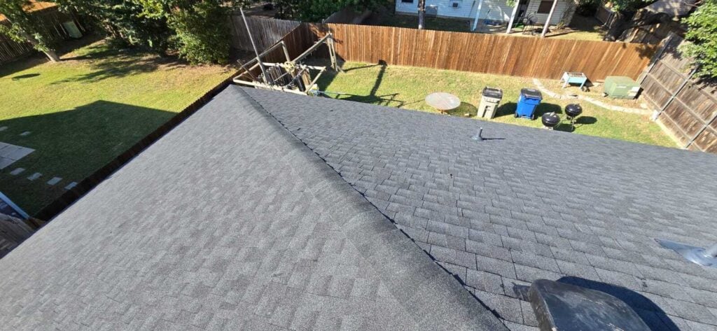 An aerial view of a residential roof covered in dark grey asphalt shingles, looking down into a backyard with a wooden fence, green grass, and outdoor items like grills and trash cans.