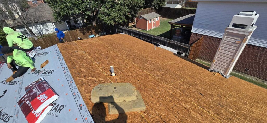 Roofers are installing underlayment on a residential roof, with two crew members visible in the foreground and one further back. The image shows the exposed roof deck, a chimney, and a vent pipe, with a backyard and shed in the background.