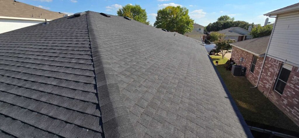 An aerial view of a residential roof covered with dark gray asphalt shingles, featuring ridge vents and a clean appearance. In the background, other houses, trees, a white van, and several people sitting on the grass next to the house are visible.