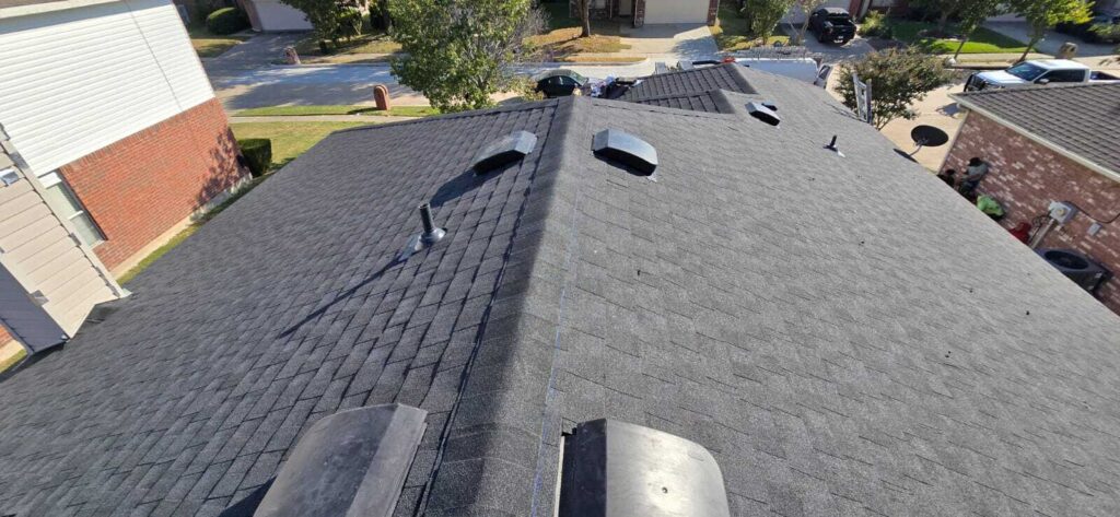 An elevated view of a dark asphalt shingle roof, showing several vents and a pipe boot. A few workers are visible on the side of the house, near an HVAC unit.