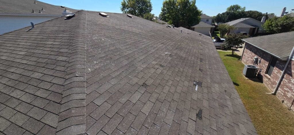An aerial view of a residential asphalt shingle roof showing significant damage, including missing shingles and exposed underlayment. Several roof vents are visible, along with a neighboring house and yard in the background.
