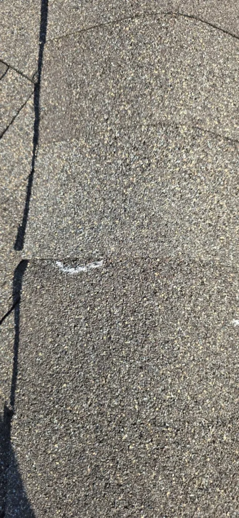 A close-up shot of asphalt shingles with a white chalk mark on one shingle, likely indicating an area of damage or concern.