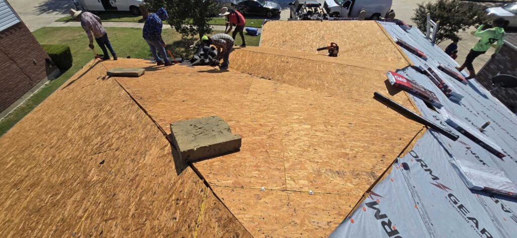 A roofing crew is actively working on a residential roof, with much of the old material removed, exposing the plywood deck. Some new underlayment is already installed, and bundles of new shingles are visible on the roof.