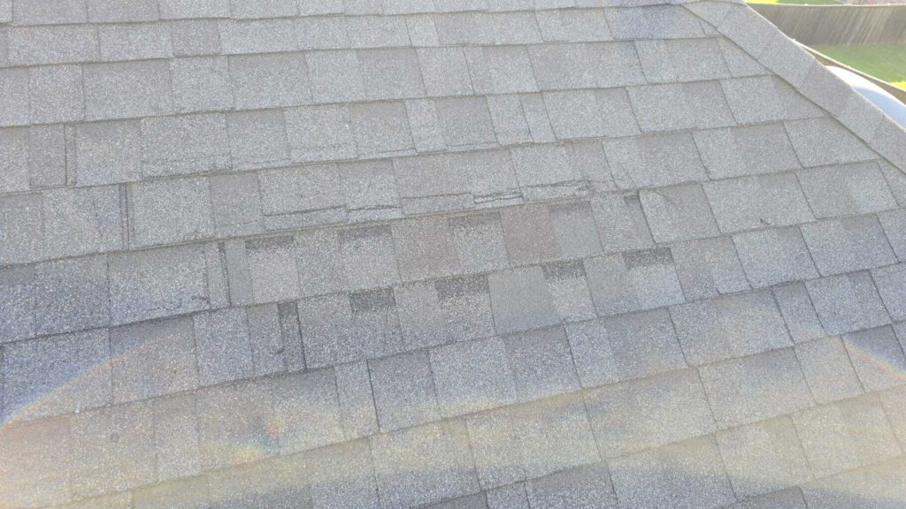 An aerial view of a gray asphalt shingle roof showing signs of wear and damage, with some shingles appearing lifted or creased. A wooden fence and green grass are visible in the background.