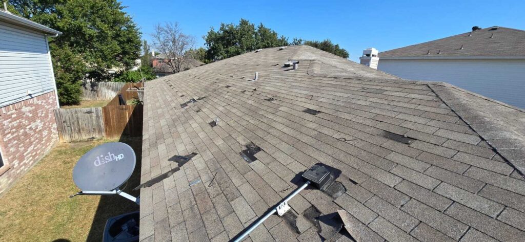 An aerial view of a residential asphalt shingle roof showing extensive damage, including missing shingles and exposed underlayment. A satellite dish is visible in the lower left foreground.