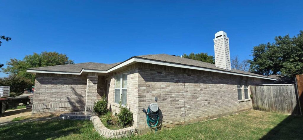 This image shows a single-story brick house with an asphalt shingle roof under a clear blue sky. A truck loaded with roofing materials is partially visible on the left side of the frame.