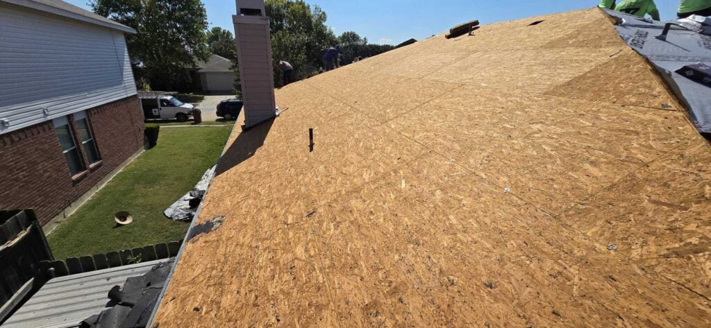 This image shows a residential roof with the old shingles removed, exposing the OSB decking. Several crew members are visible working on the roof, with some underlayment already installed.