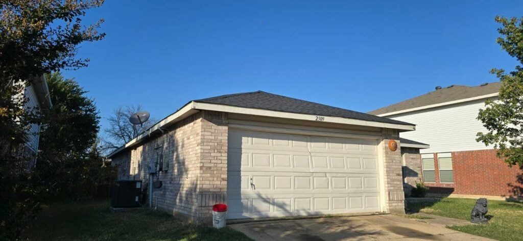 A single-story brick house with a newly installed dark asphalt shingle roof is visible under a clear blue sky. The house features a prominent garage door and a small front yard.