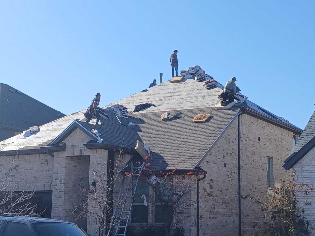 Several roofers are actively working on a residential roof, which is partially stripped down to the underlayment with new bundles of shingles ready for installation. A ladder is propped against the house, and the brick exterior is visible.