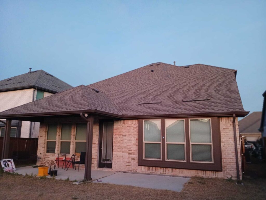 This image shows the back of a residential brick house featuring a brown asphalt shingle roof and a covered patio area with some outdoor furniture. The sky above is clear blue.