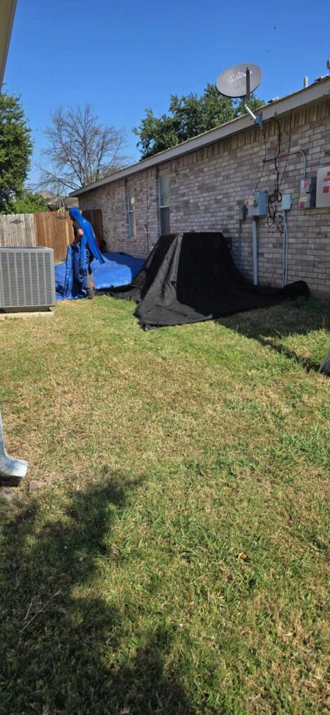 A person in blue clothing is seen in a residential backyard, handling blue and black tarps next to an AC unit and a brick house. The tarps are likely for roof protection during a job.
