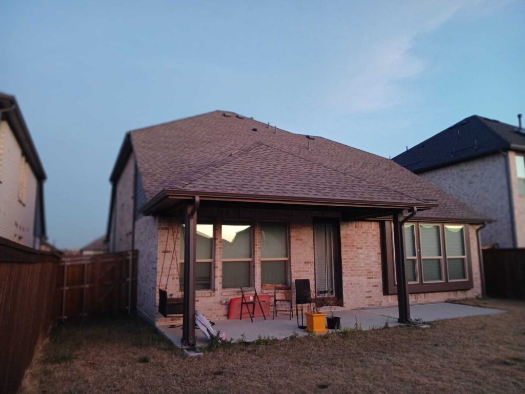 This image shows the back exterior of a brick residential house featuring a brown asphalt shingle roof and a covered patio area with some outdoor furniture. The lawn in the foreground appears dry, and parts of neighboring houses are visible on either side.