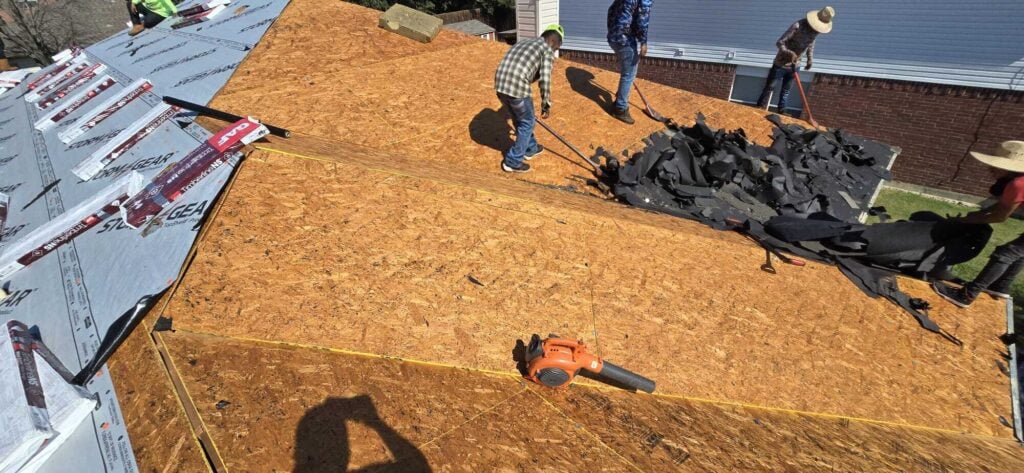 Several roofers are actively working on a residential roof, removing old asphalt shingles and preparing the roof deck. Bundles of new GAF Timberline NS shingles and underlayment are visible on one section of the roof.