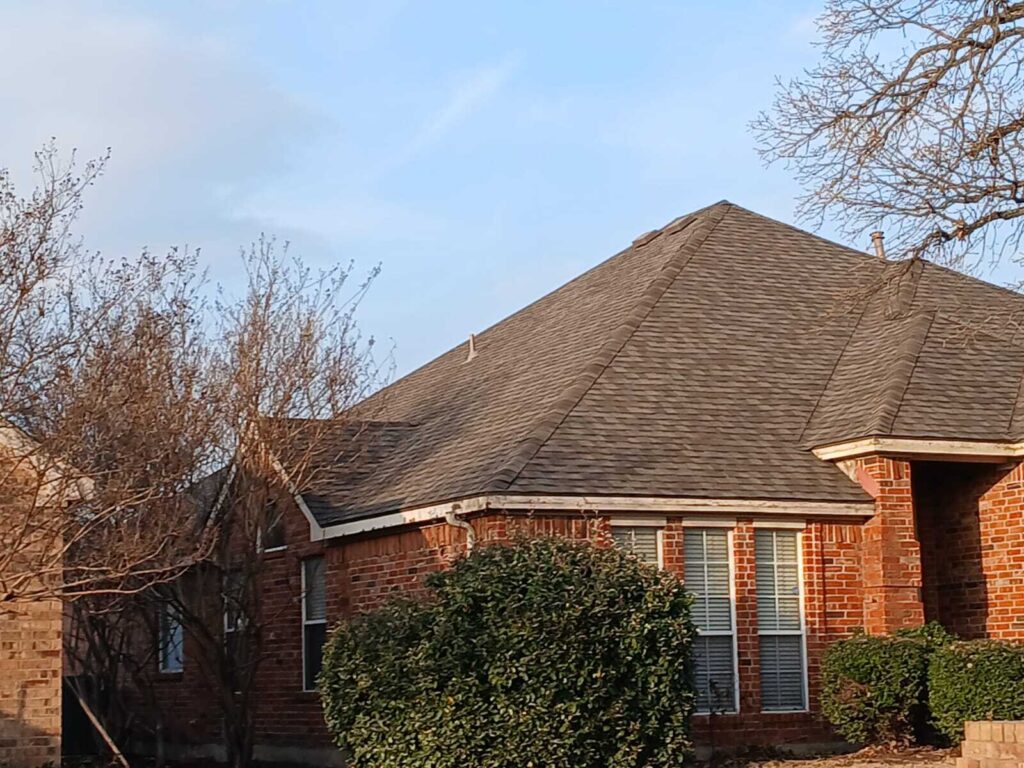 An exterior shot of a brick residential house featuring an asphalt shingle roof with visible wear and some lifted shingles. Bare trees and green bushes frame the house under a clear sky.