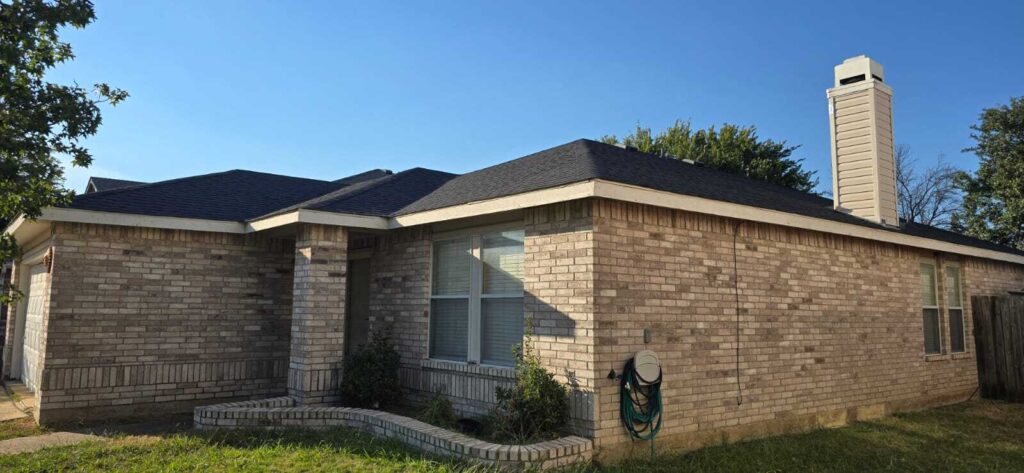 A single-story brick house with a newly installed dark asphalt shingle roof is shown under a clear blue sky. The image captures the front and side of the house, including a garage door, windows, and a chimney.
