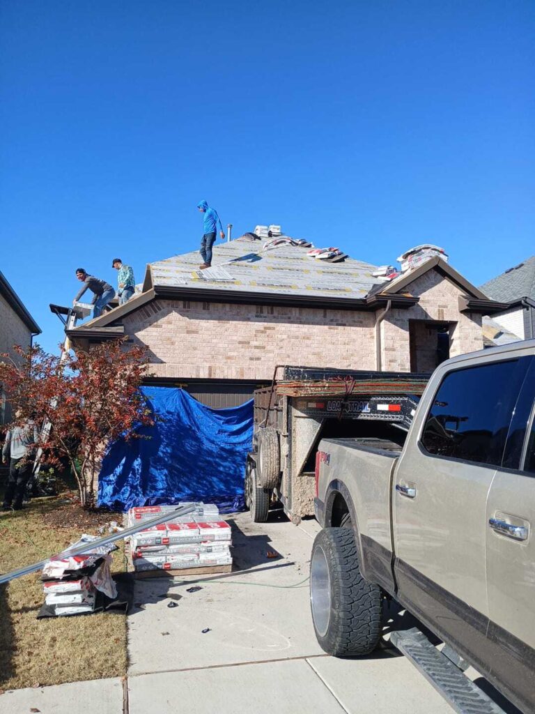 Several roofers are actively working on a residential asphalt shingle roof, with materials stacked on the driveway and a work truck and trailer parked in front of the house.