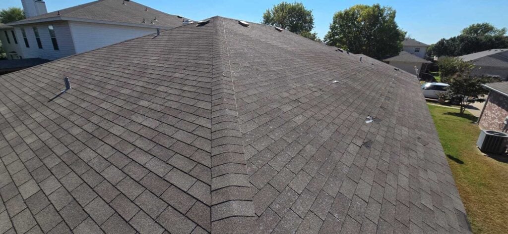 An aerial view of a residential asphalt shingle roof showing signs of wear and tear, including missing granules and some lifted shingles. Other houses and trees are visible in the background under a clear sky.