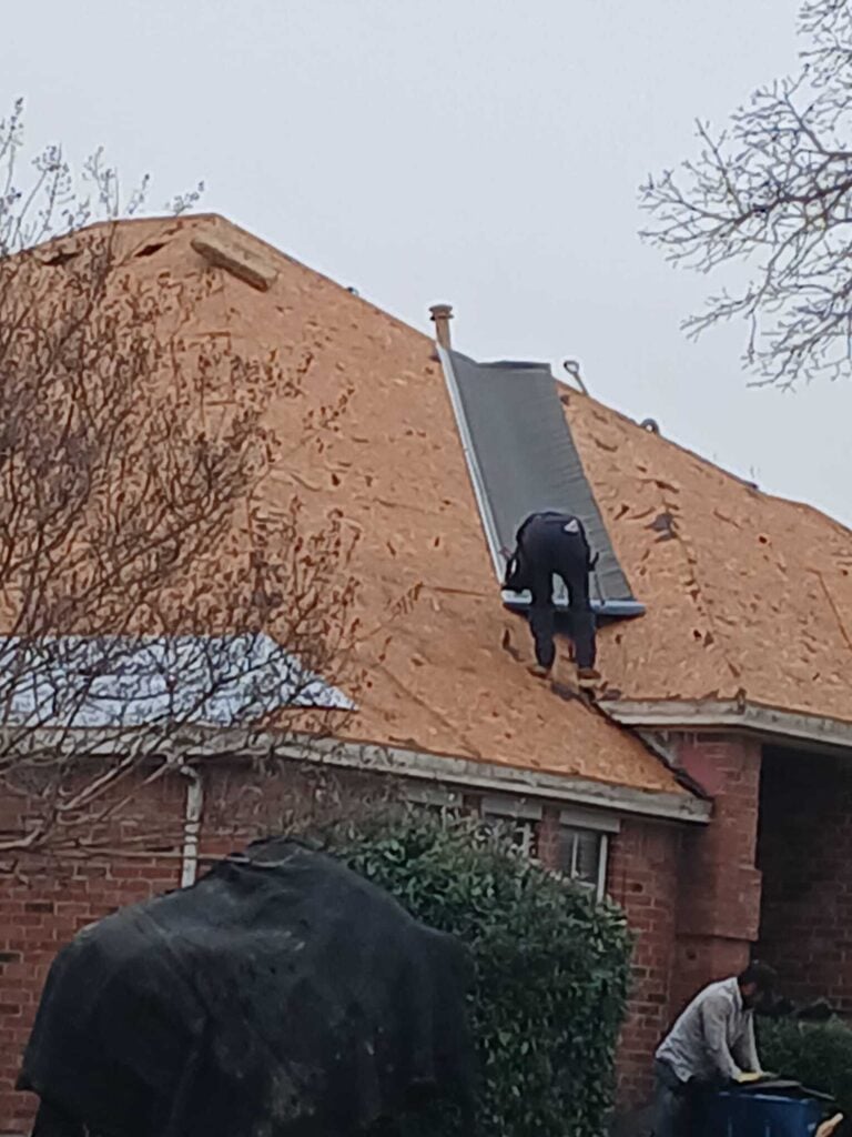 A roofer is installing underlayment on a partially stripped residential roof, while another worker is visible on the ground next to a blue bin. The house has brick siding and is surrounded by bare trees and bushes.
