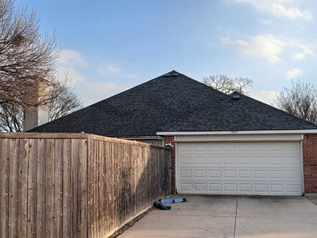 An asphalt shingle roof on a residential house, viewed from the driveway behind a wooden fence. The house features a two-car garage and brick siding.
