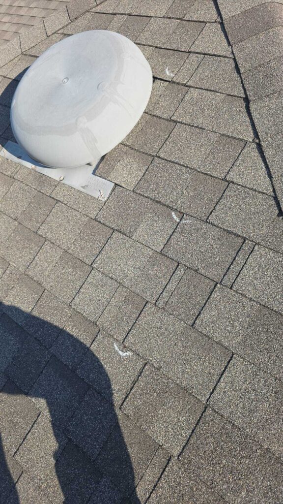 An overhead view of an asphalt shingle roof with a white satellite dish and several white chalk marks indicating potential hail damage. A shadow of a person is visible in the lower left corner.