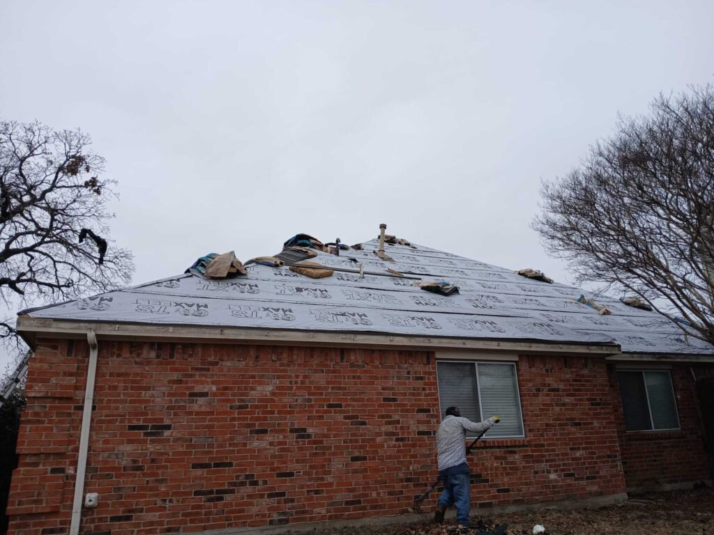 A residential house with its roof covered in Maxfelt TS underlayment, ready for new shingles. A worker is visible on the ground raking leaves next to the brick wall.