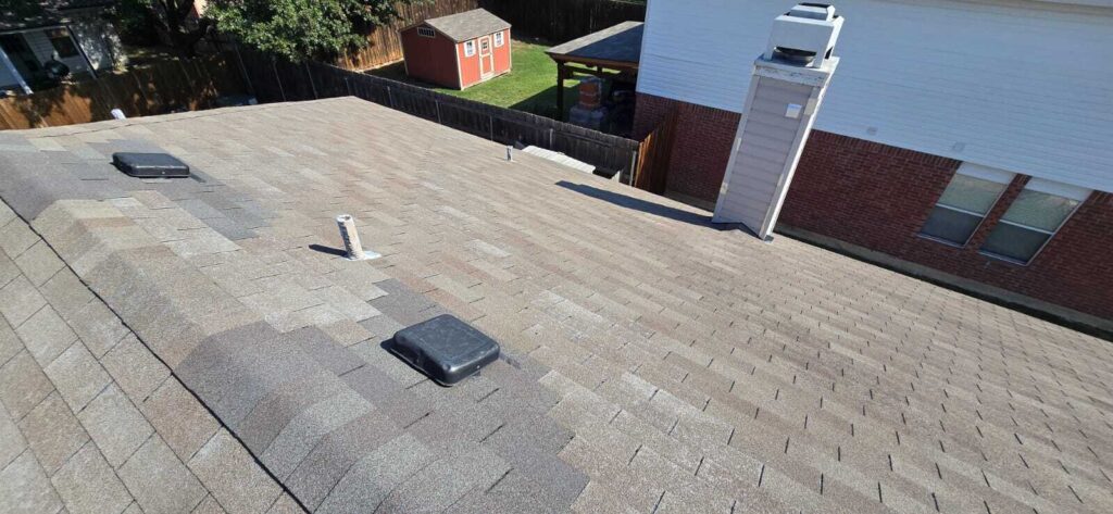 An aerial view of a residential asphalt shingle roof, featuring vents and a chimney. A red shed and a portion of the house's brick wall are visible in the background.