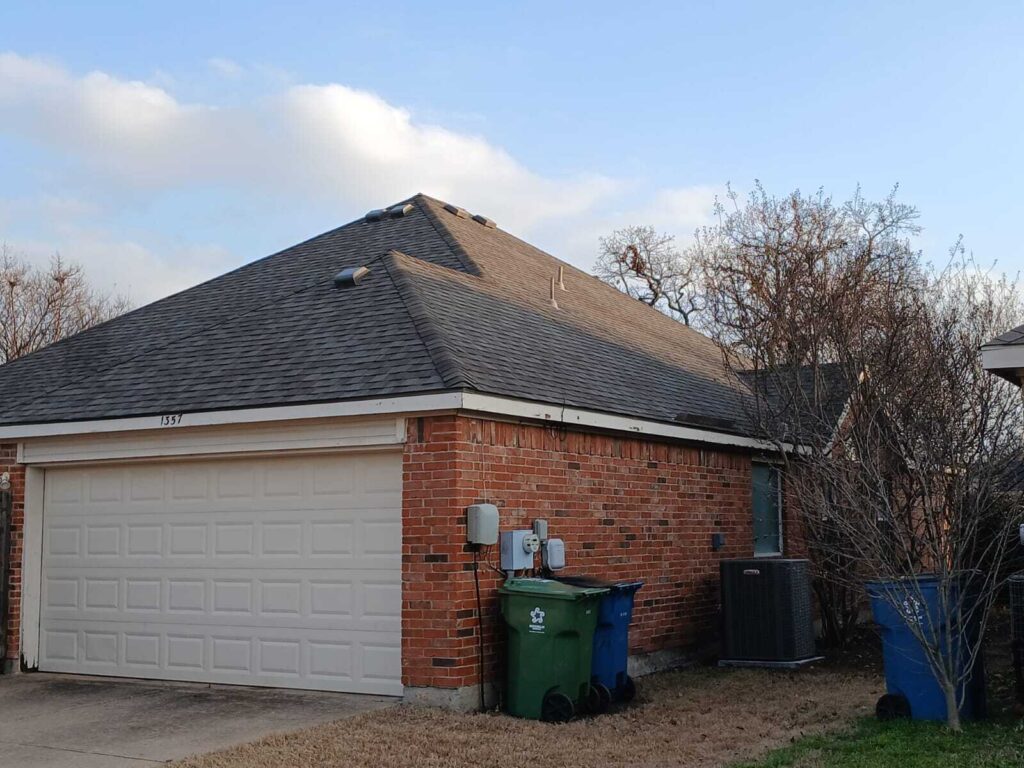 This image shows the front and side of a residential house with a dark asphalt shingle roof, brick walls, and a white garage door. Trash cans and an air conditioning unit are visible on the side of the house.