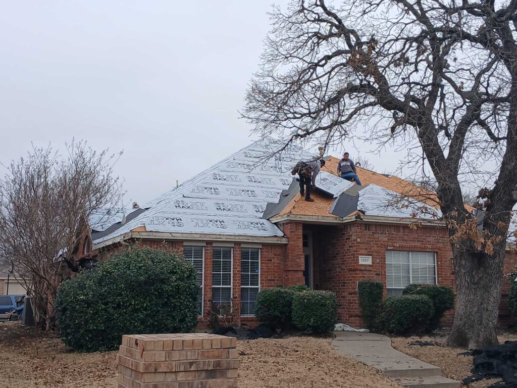 Two roofers are actively working on a residential roof, which is partially stripped and covered with underlayment. The brick house has a visible house number and is surrounded by dormant trees and shrubs under an overcast sky.