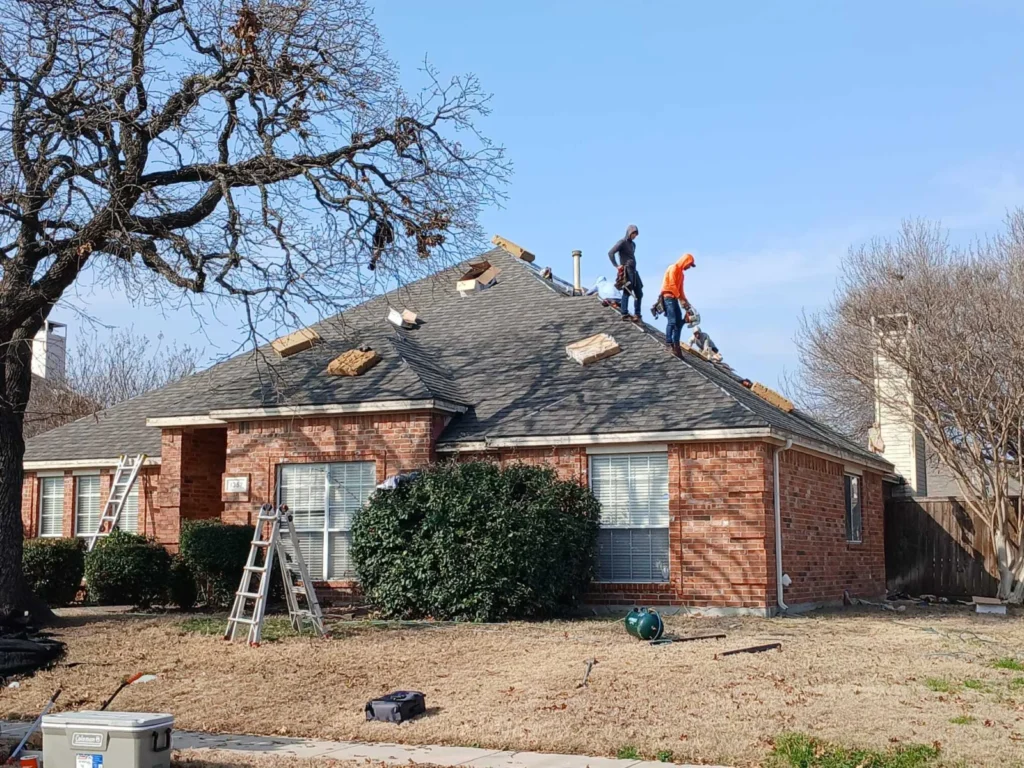 A crew is actively working on the roof of a single-story brick house, removing old asphalt shingles. Ladders and roofing tools are visible on the dry, grassy lawn.