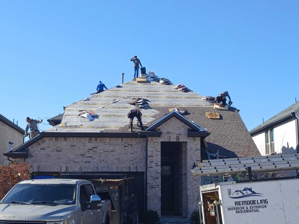 Several roofers are actively working on a residential roof, which is partially stripped and has new underlayment installed. Bundles of new asphalt shingles are visible on the roof, and a work truck is parked in the driveway.