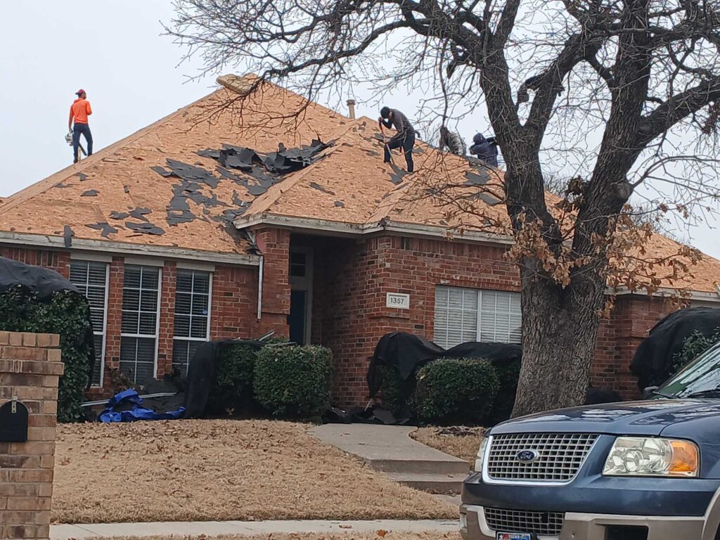 Several roofers are actively stripping old asphalt shingles from a residential house, revealing the plywood decking underneath. Protective tarps are laid around the base of the house, and a blue Ford SUV is partially visible in the foreground.