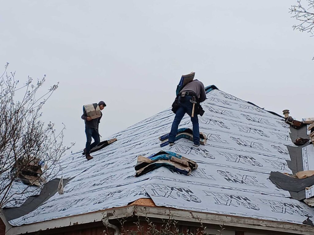 Two roofers are actively working on a residential roof, which is currently covered with underlayment. One worker is carrying a bundle of shingles, while the other is positioned near more bundles on the roof.