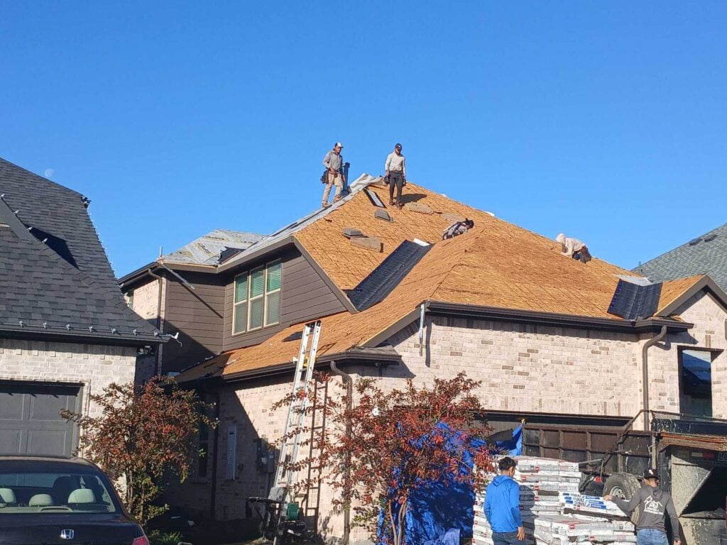 Several roofers are actively working on a residential roof, with some sections already stripped down to the decking and new shingles being installed. Bundles of new asphalt shingles are stacked on the ground next to a trailer.